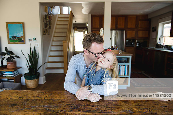 Reifer Mann sitzt am Küchentisch und küsst seine Tochter auf die Wange