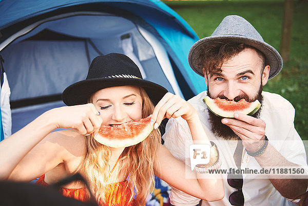 Junges Paar in Trilbies macht auf dem Festival ein Smiley-Gesicht mit Melonenscheibe