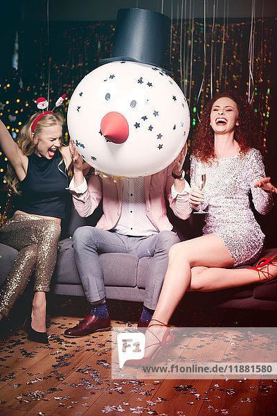 Friends sitting together at party  man holding snowman head in front of face  women laughing