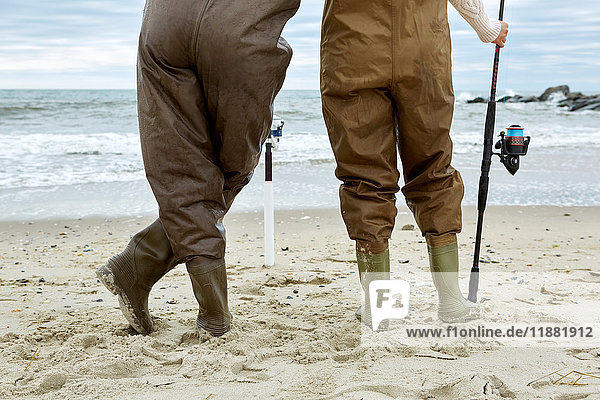 Waist down rear view of young couple in sea fishing waders on beach