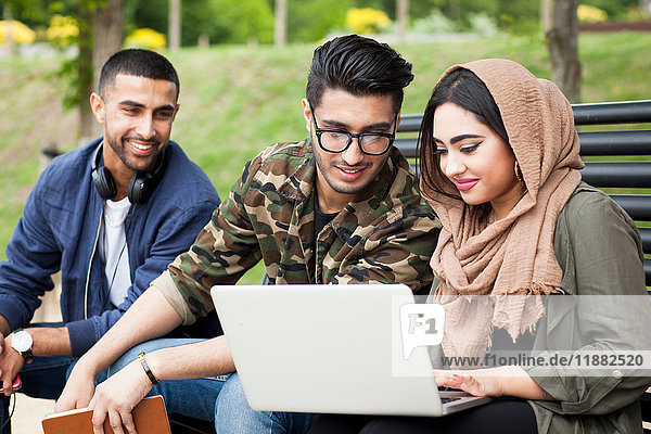 Drei Freunde  die auf einer Parkbank sitzen und auf einen Laptop schauen