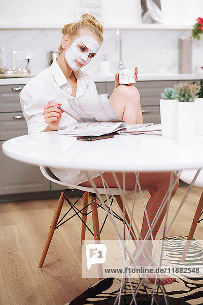 Frau mit Gesichtsmaske sitzt am Esstisch und liest Zeitschrift