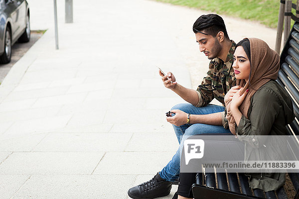 Young couple sitting on park bench  young man looking at smartphone