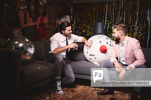 Two men sitting on sofa at party  giant snowman head in-between them  laughing
