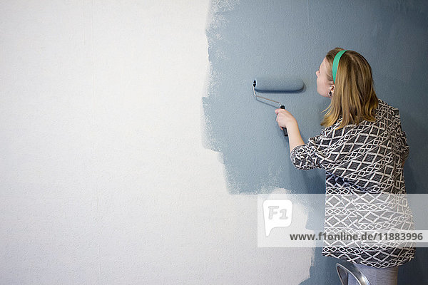 Junge Frau auf Trittleiter trägt zu Hause graue Farbe auf Innenwand auf