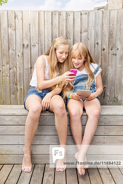 Im Garten sitzende Mädchen mit digitalem Tablet und Smartphone lächeln
