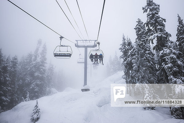 Abfahrtskifahrer fahren mit dem Sessellift in einem Skigebiet bei Schneetreiben und Wolken; Whistler  British Columbia  Kanada'.