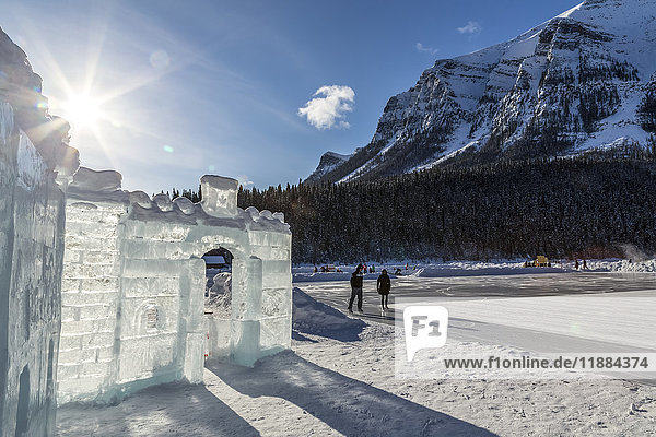 Die Sonne scheint hinter einer Eisburg  die auf dem zugefrorenen Lake Louise im Banff National Park errichtet wurde  während im Hintergrund Eiskunstläufer auf dem See laufen; Lake Louise  Alberta  Kanada'.