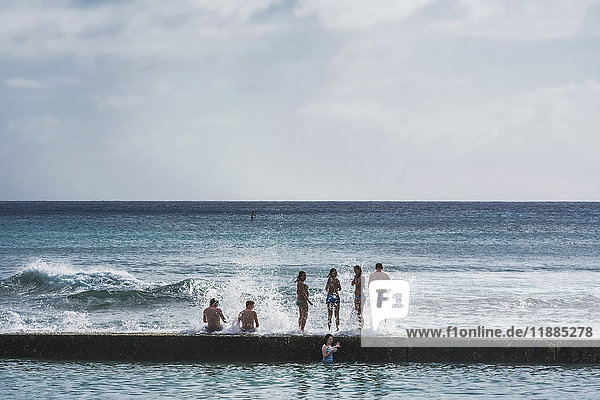 Touristen  die auf einer Mauer sitzen und stehen  während die Wellen mit Blick auf den Ozean hereinplatschen; Honolulu  Oahu  Hawaii  Vereinigte Staaten von Amerika'.