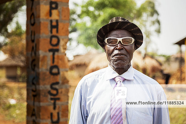 Ein Mann mit weißer Brille posiert vor einem Fotostudio; Uganda'.