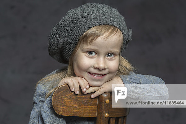 Porträt eines lächelnden Mädchens mit Strickmütze auf einem Holzstuhl vor grauem Hintergrund.