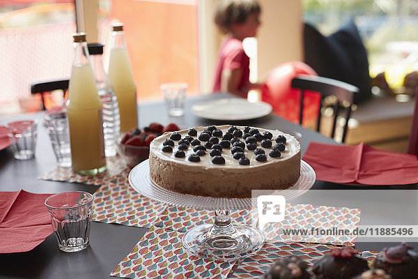 Torte auf Ständer über Platzmatten durch Flaschen am Tisch mit spielendem Mädchen im Hintergrund
