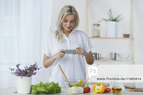 Lächelnde junge Frau fotografiert frischen Salat durch Smartphone in der Küche