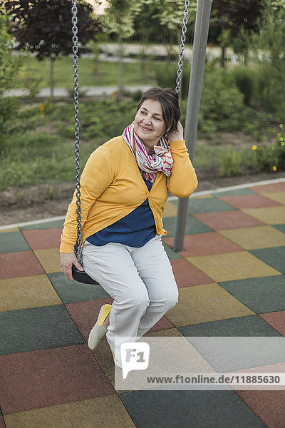 Lächelnde Frau schaut weg,  während sie auf der Schaukel auf dem Spielplatz sitzt.