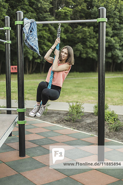 Fröhliche junge Frau hängt am Seil der Kletterhalle im Park.