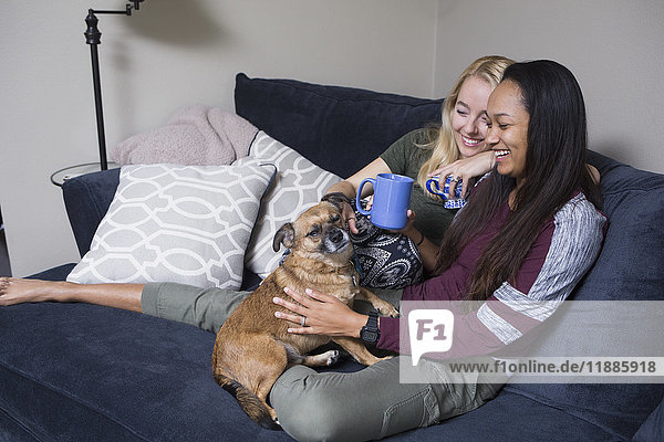 Happy young women sitting with coffee cups and dog on sofa at home