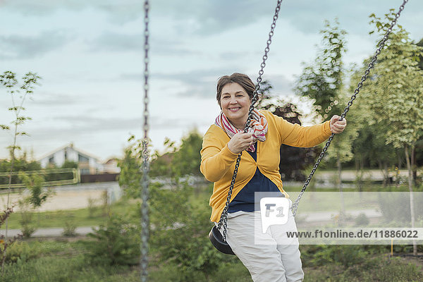 Lächelnde Frau spielt auf Schaukel auf dem Spielplatz gegen den Himmel