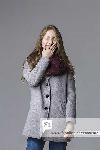 Smiling teenage girl covering mouth standing against gray background
