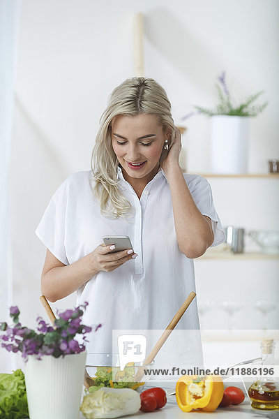 Smiling young woman using mobile phone while preparing salad at kitchen counter