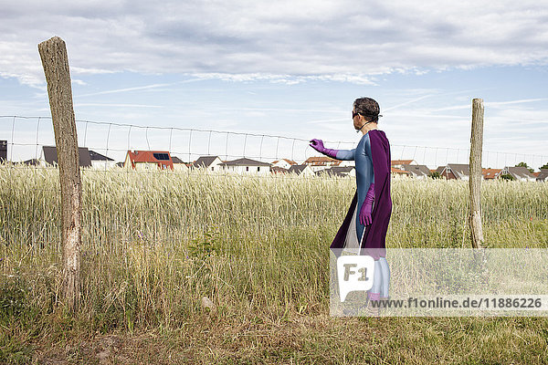 Mann im Superheldenkostüm steht am Zaun auf grasbewachsenem Feld gegen den Himmel.