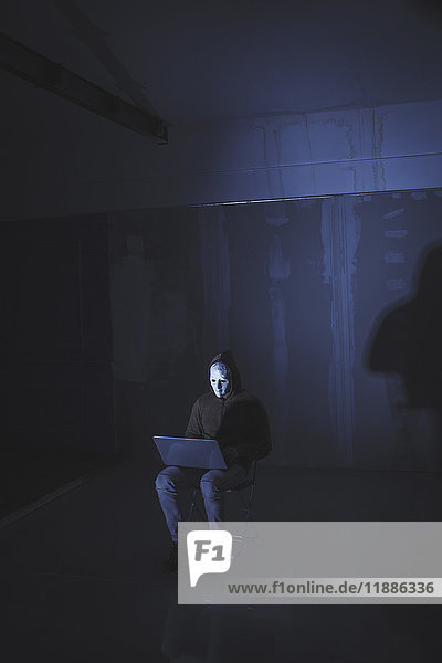 Full length of computer hacker wearing mask using laptop while sitting against wall