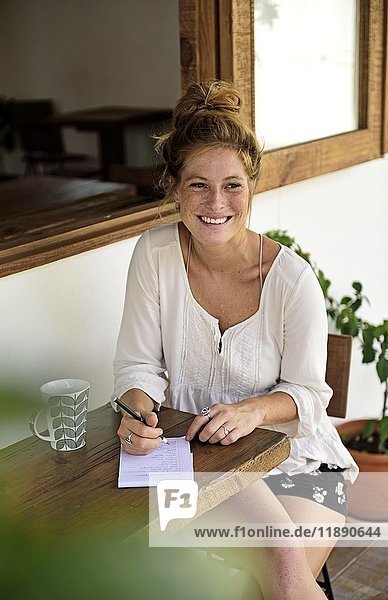 Portrait of laughing young woman with cup of coffee sitting on veranda of a coffee shop writing