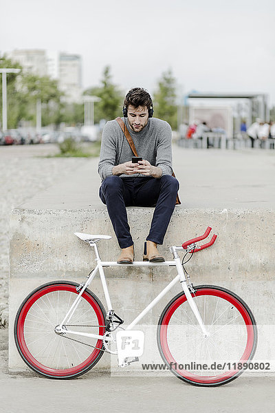 Young man with racing cycle sitting on a wall listening music with headphones