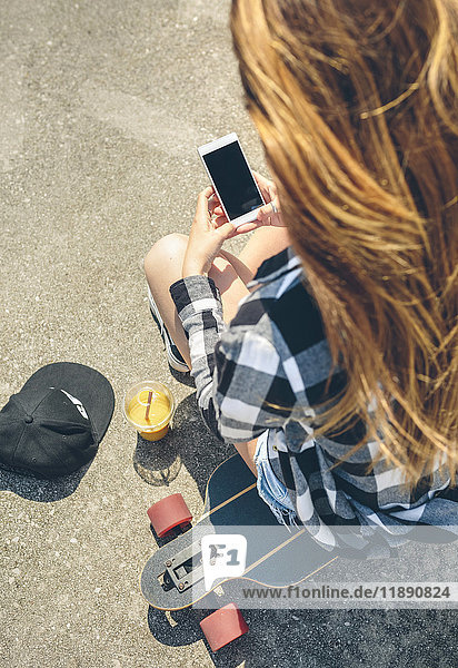 Rückansicht einer jungen Frau mit langen Haaren  die auf dem Longboard sitzt und auf das Smartphone schaut.