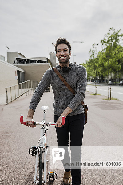Porträt eines lächelnden jungen Mannes mit Kopfhörer  der sein Fahrrad schiebt.