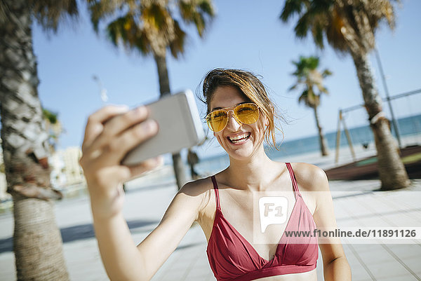 Lachende junge Frau mit gelber Sonnenbrille  die einen Selfie auf der Strandpromenade trägt.