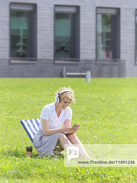 Woman with headphones sitting on a meadow looking at cell phone