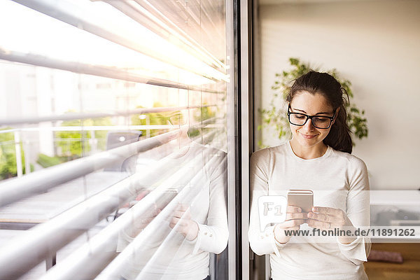 Lächelnde Frau steht am Fenster zu Hause mit dem Smartphone