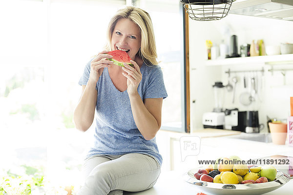 Reife Frau  die in der Küche sitzt und Wassermelone isst.