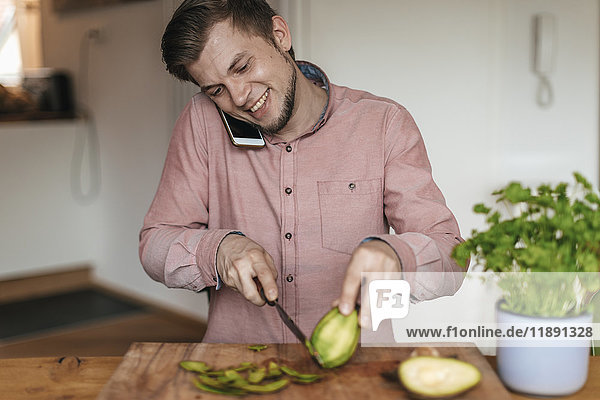 Smiling man on the phone chopping acocado in the kitchen