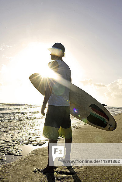 Mann mit Surfbrett steht am Strand und beobachtet den Sonnenuntergang.