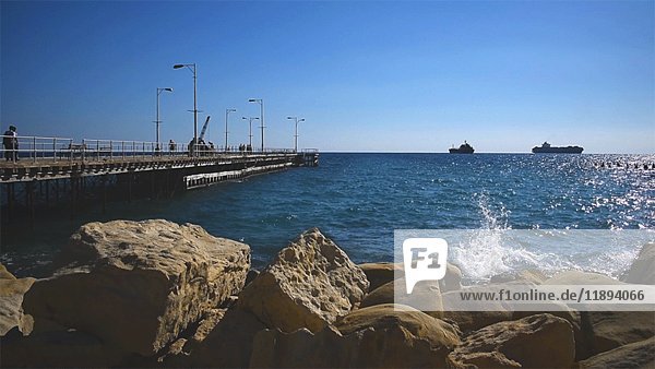 Wellen plätschern gegen Felsen in der Nähe von Pier mit zwei Booten am Horizont im Hintergrund  Limassol  Zypern