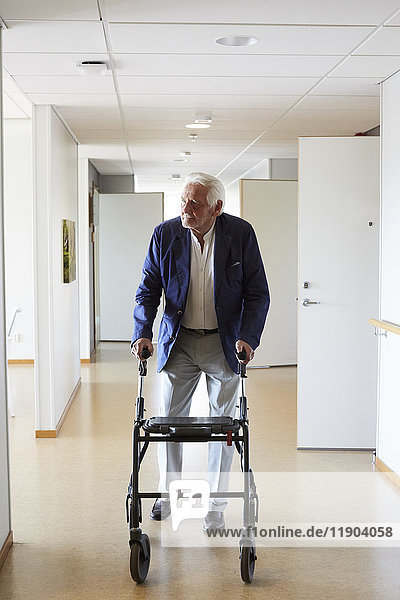 Senior man looking away while walking with rollator in hospital corridor