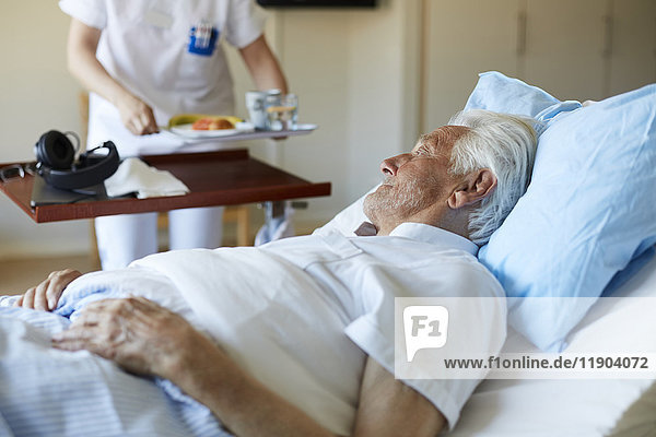 Älterer Mann schaut auf eine Krankenschwester  die das Frühstück serviert  während sie auf dem Krankenhausbett liegt.