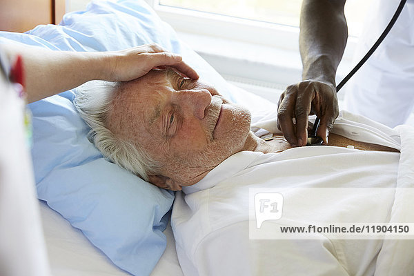 Abgeschnittenes Bild einer Krankenschwester  die den älteren Mann tröstet  während ein männlicher Kollege mit einem Stethoskop auf der Krankenstation untersucht.