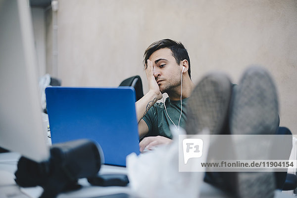 Tired computer programmer using laptop while sitting with feet up at desk in office