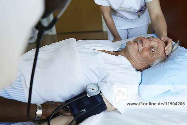 Krankenpfleger  der den Blutdruck eines älteren Mannes auf dem Krankenhausbett überprüft  während ein Kollege den Patienten trösten kann.