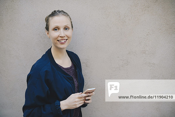Porträt einer glücklichen Computerprogrammiererin  die ein Smartphone hält  während sie an der beigen Wand im Büro steht.