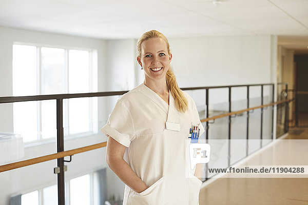 Porträt einer lächelnden Krankenschwester  die mit den Händen in den Taschen am Krankenhausflur steht.