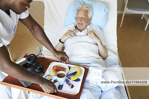 Hochwinkelansicht des Krankenpflegers  der das Frühstück für den älteren Mann auf der Krankenstation serviert.