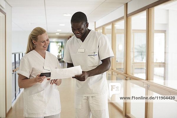 Glückliche Krankenschwester und Krankenpflegerin mit digitalem Tablett bei der Untersuchung von Dokumenten im Krankenhauskorridor