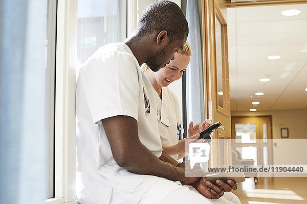 Multiethnische Krankenschwestern mit Smartphone auf der Fensterbank im Krankenhaus
