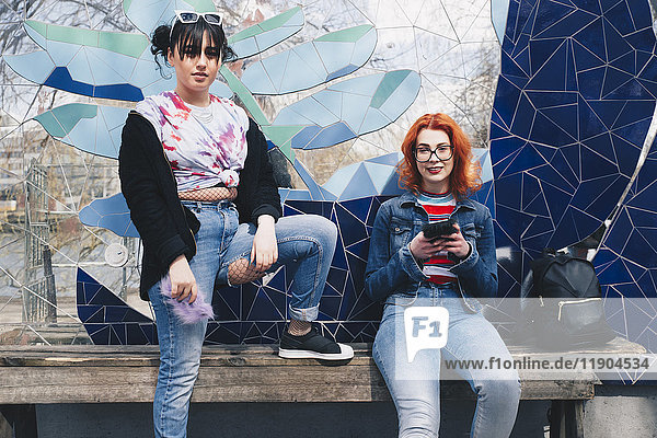 Porträt von Hipster-Freundinnen,  die Mobiltelefone an der Mosaikwand halten