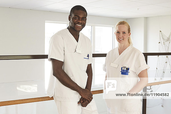 Portrait of happy male and female nurses standing against railing at hospital corridor