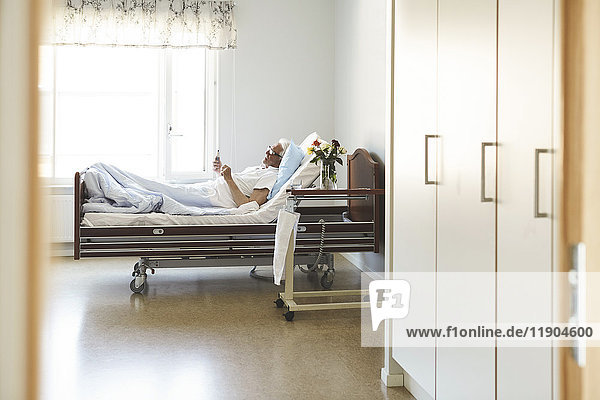 Seitenansicht eines älteren Mannes  der ein Smartphone benutzt  während er auf dem Bett in der Krankenstation liegt.