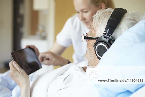 Senior-Mann mit Kopfhörer bei der Benutzung des digitalen Tabletts mit Krankenschwester auf dem Krankenhausbett